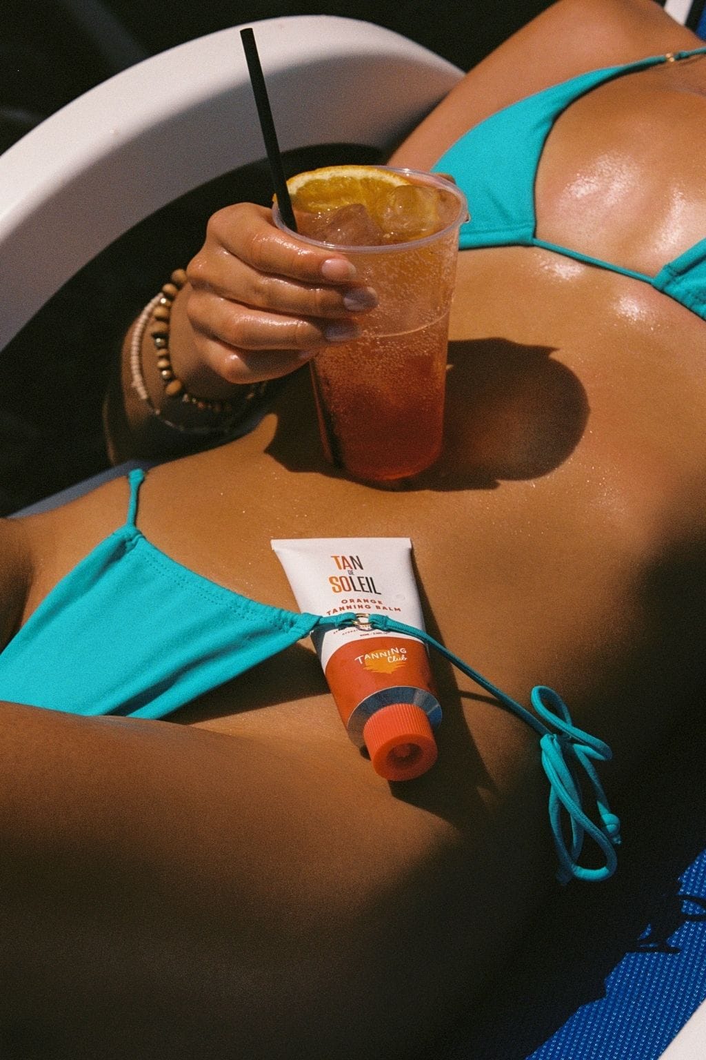 Person in turquoise bikini holding beverage with Tan de Soleil tanning products displayed on tanned skin.
