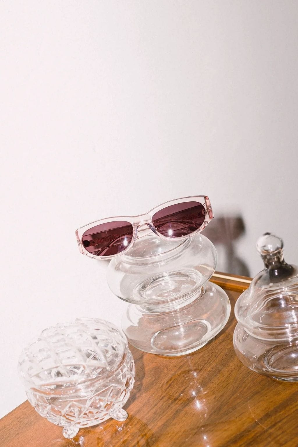 Crystal rose-tinted oval sunglasses with plum-colored lenses resting on stacked clear glass bowls.