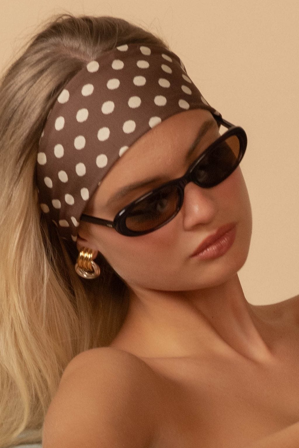 Brown headband with white polka dots worn on head above dark oval sunglasses.