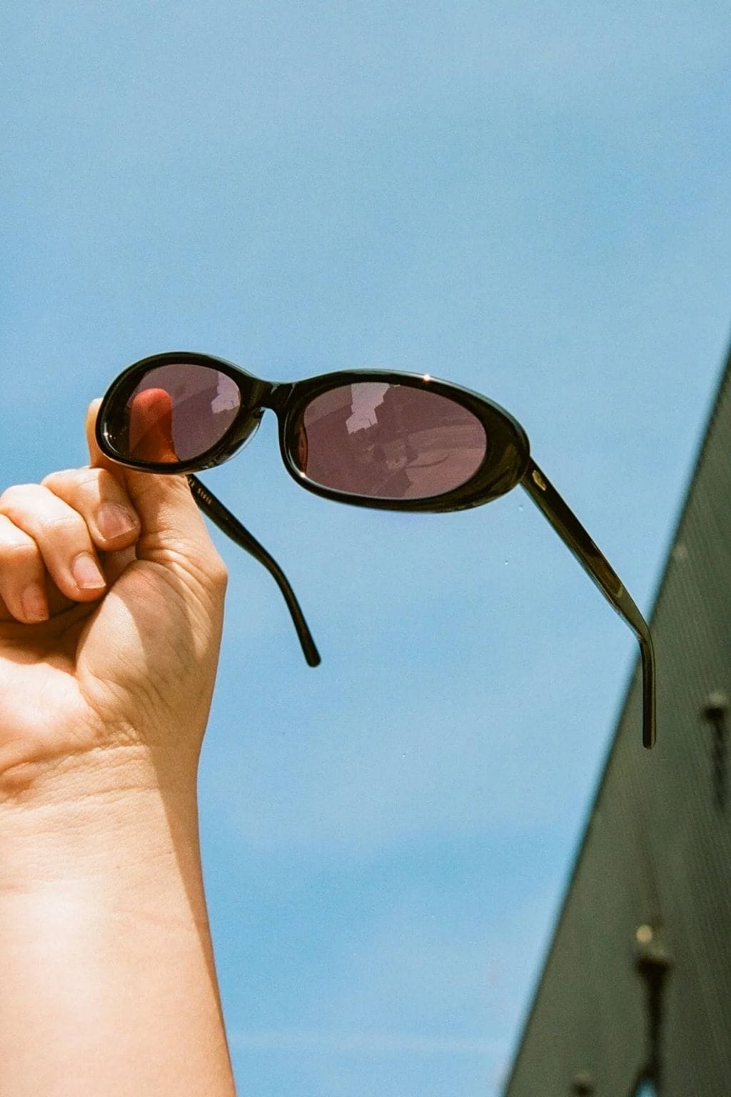Black oval sunglasses with dark tinted lenses held in a hand against a blue sky.