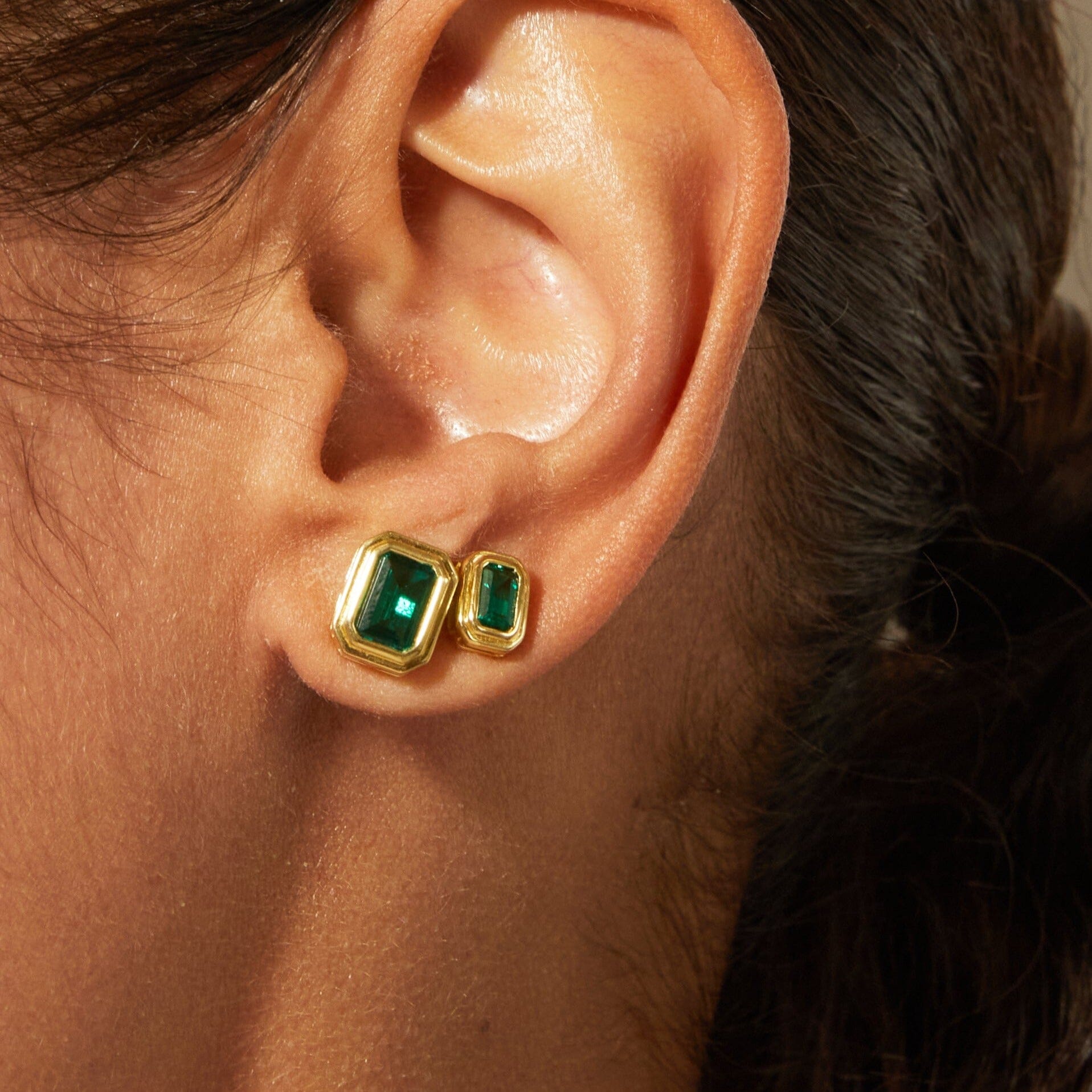 Pair of gold-framed stud earrings with rectangular green emerald stones worn on earlobe.