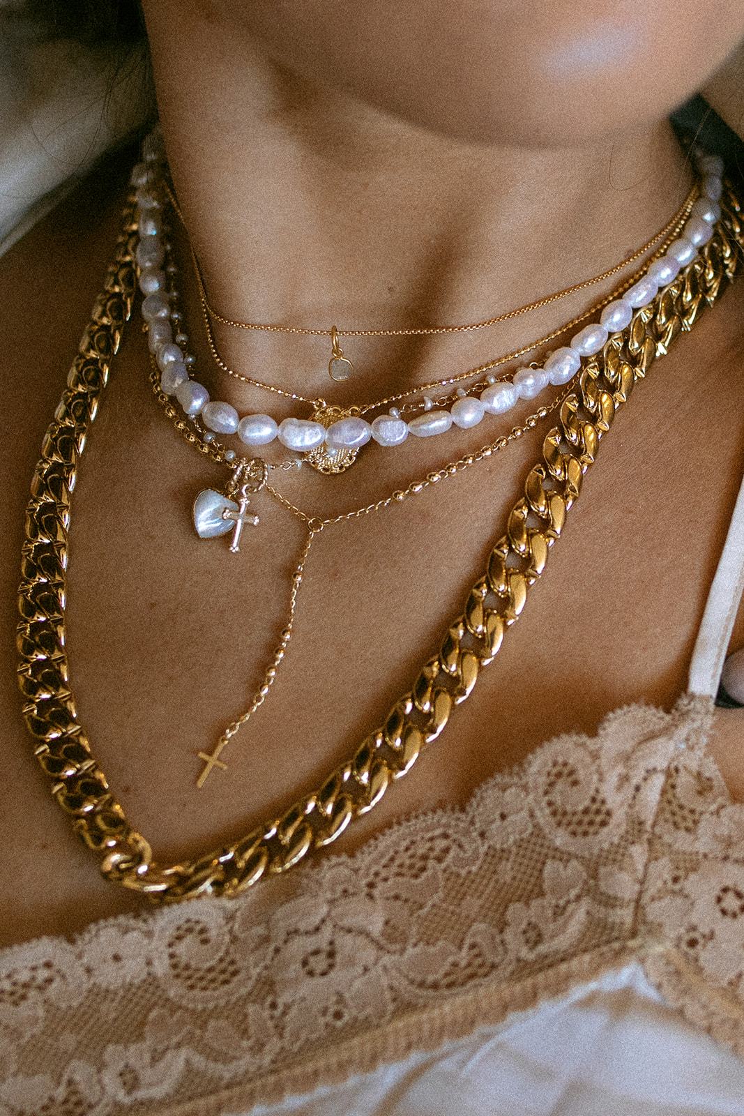 Layered gold chain necklace with white pearl beads, cross pendant, and pearl charm worn on model's neck.