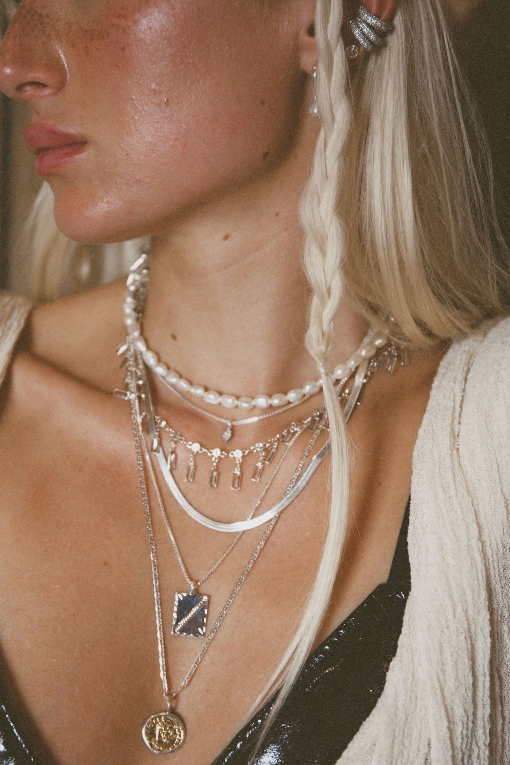 Woman wearing a layered necklace with pearls, silver chains, and pendant charms including a rectangular and circular design.