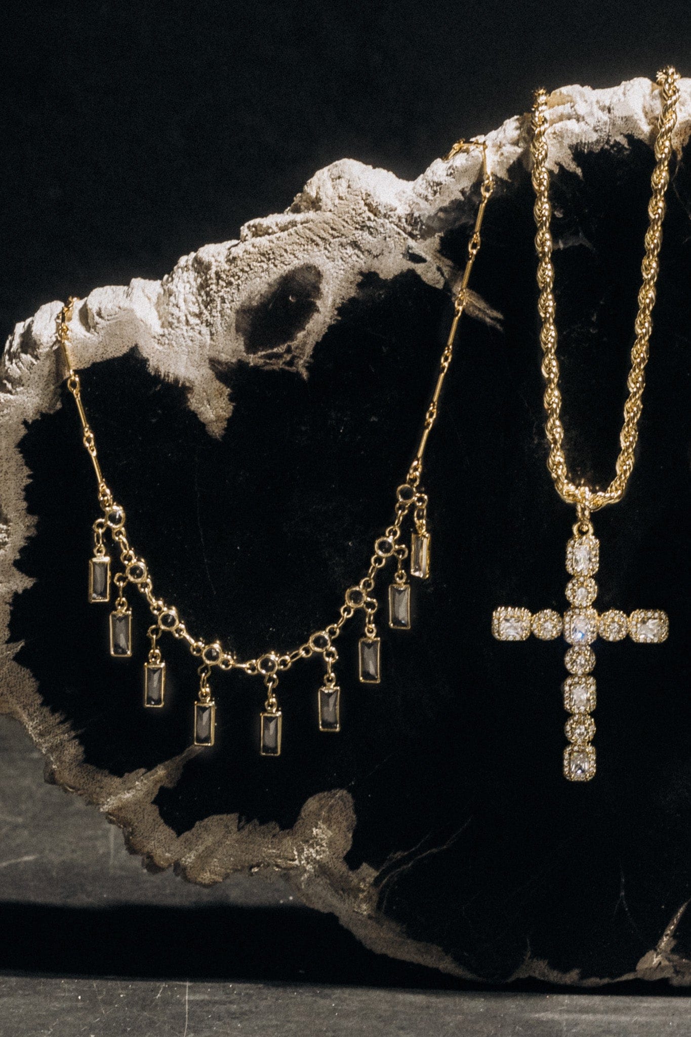 Gold-toned necklaces with cubic zirconia rectangular and cross-shaped pendants displayed on a white textured branch against b